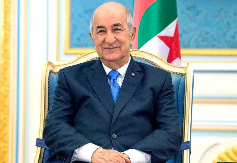 Image du président algérien Abdelmadjid Tebboune assis et entrain de sourire à côté du drapeau d’Algérie